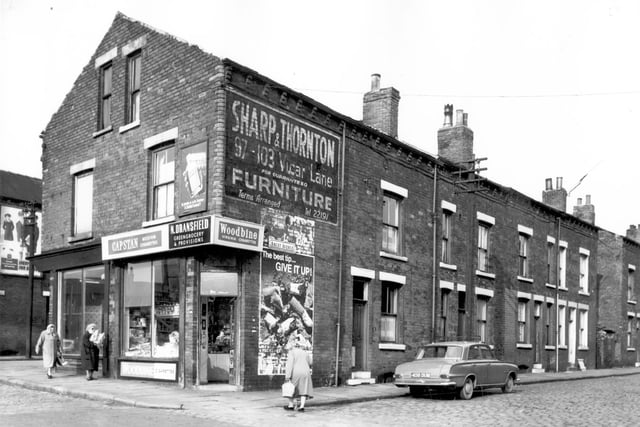 A shop at junction of Ascot Avenue and Pontefract Lane. Two women walk towards the N. Dransfield greengrocery & provisions store from the left, while another woman crosses the street on the right to reach the store. Above the shop are two advertisements for Dettol on the left and Sharp & Thornton furniture store on the right. On the right a row of back-to-back terraces are visible with a car, reg: 408 DUM parked outside. Pictured in October 1966.
