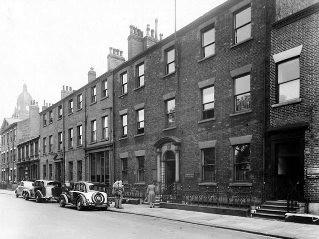 Three people wait outside The Register Office on Park Square East in July 1949. Above the buildings, the clock of Leeds Town Hall is visible (far left).