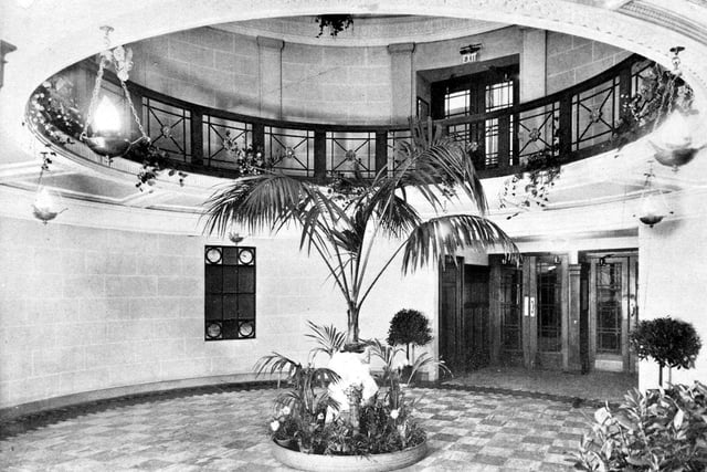 Inside the foyer of the new Majestic Cinema on City Square. Trailing plants and lamps hang from the circular balcony. A circular planter contains exotic tropical plants including a tall Parlour Palm as a centrepiece.
