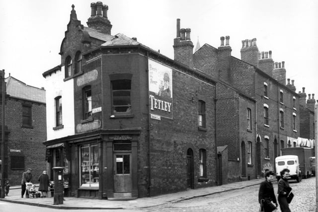 On the left are two commercial properties on Tong Road. Painted white, is a second hand furniture shop where a man, woman and child inspect the goods on display outside. Fred Brown's off-licence and tobacconists follows. A large, painted sign for Tetley beer is visible on the wall. Dewhirst Place runs to the right edge where two women cross the road. Pictured in May 1965.