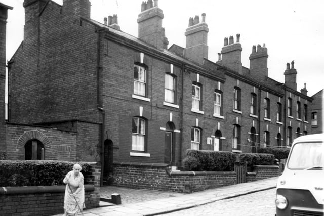 Long view of back-to-back terraced houses on Algeria Street in July 1964. Far left an archway leads into the yard where the outside toilets are situated for this section of the street. Right of that another arched entrance defines a passage which cuts through to Broadway Street. A lady sweeps the footpath close to her house.