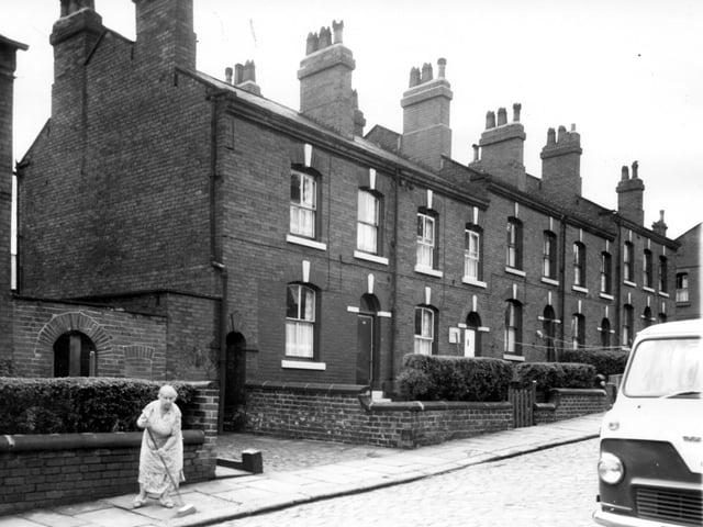 Long view of back-to-back terraced houses on Algeria Street in July 1964. Far left an archway leads into the yard where the outside toilets are situated for this section of the street. Right of that another arched entrance defines a passage which cuts through to Broadway Street. A lady sweeps the footpath close to her house.