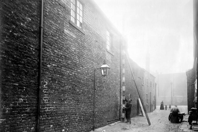 Mabgate Green in September 1911. Brick built commercial building with electric street light fixed to wall. Two workmen, one up ladder, measure building height with pole. Another workman sits on box over handcart. Woman and children look on.