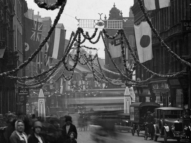 This view looks along Albion Street which has been decked with garlands and flags to celebrate Civic Week. Trellis panels are suspended over the street and are surrounded by flags including the national flags of Germany, France, Japan and England.