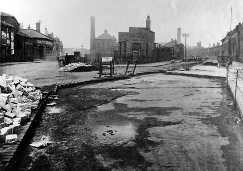 Globe Road Bridge in August 1926. Hide and skin market on the street to the left. Large part of Globe Road has been dug up. Two signs marked 'no road', block access down the road. Further down Globe Road, a group of eight workers are using pickaxes to dig the road up. Simingtons Ltd, cardboard box manufacturers is to the right of them.