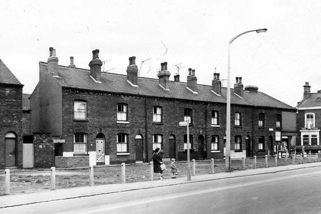 A long view of Dewsbury Terrace which is set well back from Hunslet Hall Road (foreground). The shop on the corner is number 106 Dewsbury Road and the Junction Pub can be seen far right. A woman and small boy wait for a bus on Hunslet Hall Road.