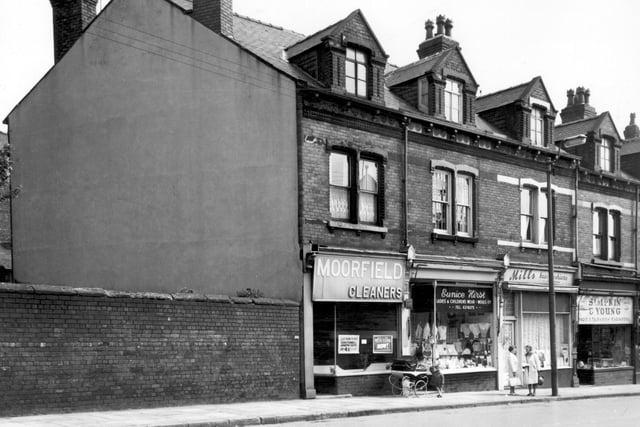 Pictured in June 1965, from left, is Moorfield Cleaners with a sign in the window which states 'Ladies Bargains this week! Costumes and Jumper Suits only 4/11d' (just under 25p). Costumes refer to a ladies skirt and jacketed suit, while a jumper suit was a knitted top and skirt. Moving right, a child holds onto a pram outside the entrance to Eunice Hirst's Ladies and Childrenswear. The window is crammed with women's tops and jumpers and girl's dresses. Two women chat outside Mills Hair Fashions and on the right is Simpkin and Young, radio and television engineers.