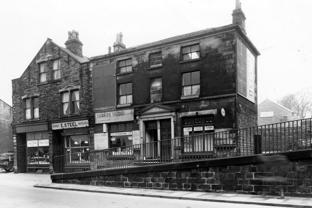 A parade of businesses in Lower Town Street in the area of Stocks Hill in March 1960. From left to right they are number 145, Williamson's high class fruiterers; number 145a, E. Steel, family butchers, and number 147, Dunkirk Studio, a photographic business.  A more recent building, the Kingdom Hall of Jehovah's Witnesses is visible in the background, right. The wall in the foreground is Grade II listed and dates from the late 18th century. It acts as a retainer to the raised pavement and is approximately 3 metres at its highest point and 30 metres in length.