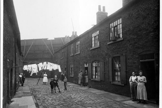 There are two lines of washing hanging across Chadwick Court and several men, women and children watching the photographer.