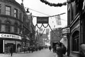 A view looks across from the end of Upperhead Row onto Guildford Street. The street decorated with garlands and flags for the Civic Week celebrations. On the left are Campbells House Furnishers and Shiphams clothiers at the entrance to the Victoria Arcade. The Guildford Hotel can be seen moving back with the Nag's Head visible on the right edge.