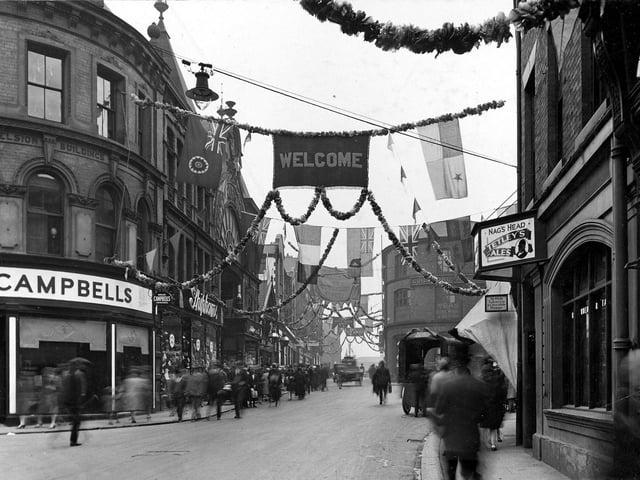 A view looks across from the end of Upperhead Row onto Guildford Street. The street decorated with garlands and flags for the Civic Week celebrations. On the left are Campbells House Furnishers and Shiphams clothiers at the entrance to the Victoria Arcade. The Guildford Hotel can be seen moving back with the Nag's Head visible on the right edge.