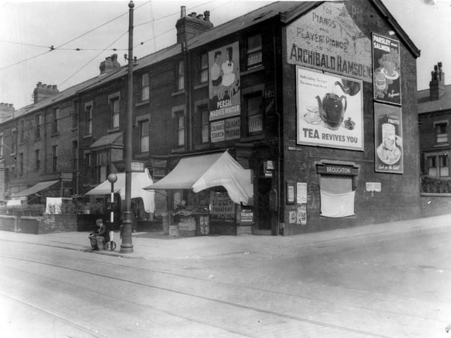 Shops and houses on Compton Road in June 1939 prior to improvement. Shops have awnings up. End shops have adverts for Persil, Colemans starch, Colemans mustard, Archibald Ramsden's pianos and others not identified. A man is sitting on the edge of the road by a belisha beacon.