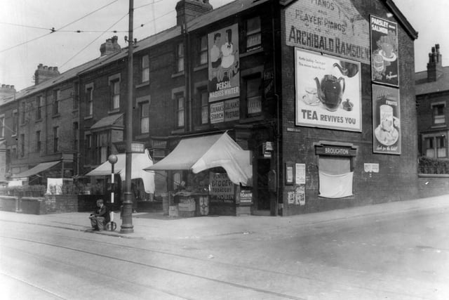 Shops and houses on Compton Road in June 1939 prior to improvement. Shops have awnings up. End shops have adverts for Persil, Colemans starch, Colemans mustard, Archibald Ramsden's pianos and others not identified. A man is sitting on the edge of the road by a belisha beacon.