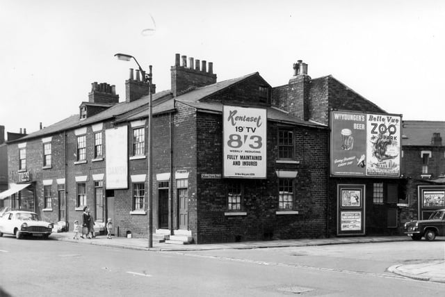 The junction of Dewsbury Road and Moor View Road in August 1964. On the far left is a Bread Shop at number 146. Also on the left are back-to-back houses with an advertising hoarding on the wall promoting Danish bacon. Two women and two children walk past a parked car towards the open door of number 140. On the right is Moor View Road displaying multiple advertisements for Rent a set 19" TV for 8'3, Wm Youngers Beer and Belle Vue Zoo Park. smaller posters promote the Elvis film Kissin' Cousins at the ABC Cinema Vicar Lane and recitals at Temple Newsam.