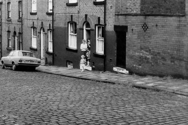 A row of double fronted back-to-back terraced houses on St Luke's Terrace with a shared outside toilet yard on the right. Two adults and two children stand in the doorway of number 7 smiling. Pictured in June 1973.