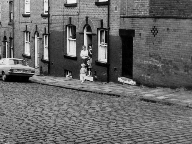 A row of double fronted back-to-back terraced houses on St Luke's Terrace with a shared outside toilet yard on the right. Two adults and two children stand in the doorway of number 7 smiling. Pictured in June 1973.