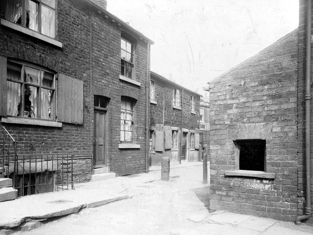 Delph Street on Woodhouse Cliff in June 1913. This is a view of an early Victorian street built in both brick and stone. Most of the ground floor windows have wooden shutters. The street is paved with rough stone blocks and there are two stone bollards visible. The opening in the lean-to type extension is to a midden for rubbish.