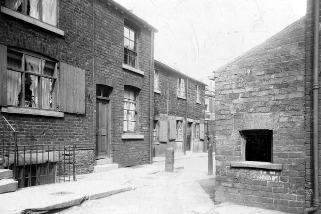 Delph Street on Woodhouse Cliff in June 1913. This is a view of an early Victorian street built in both brick and stone. Most of the ground floor windows have wooden shutters. The street is paved with rough stone blocks and there are two stone bollards visible. The opening in the lean-to type extension is to a midden for rubbish.