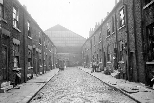 Brick built houses on Mason Street in state of decay, with broken windows, worn steps and uneven paving. Street ends in brick wall, over looked by building of former Brunswick Brewery. Row of outside lavatories face down the street. Babies pram in road. Child on left. Right, people in doorways, man stands next to a barrow, made from two pram wheels and a wooden box with two handles. Two large wooden trapdoors in pavement Pictured in August 1935.