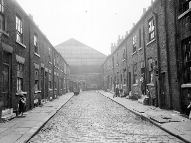 Brick built houses on Mason Street in state of decay, with broken windows, worn steps and uneven paving. Street ends in brick wall, over looked by building of former Brunswick Brewery. Row of outside lavatories face down the street. Babies pram in road. Child on left. Right, people in doorways, man stands next to a barrow, made from two pram wheels and a wooden box with two handles. Two large wooden trapdoors in pavement Pictured in August 1935.