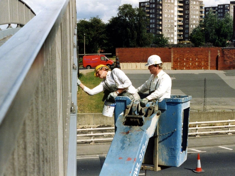 Two workmen on an access platform are painting the footbridge over the York Road flyover. The footbridge crosses the road from Quarry House, the Department of Health and Social Security, (under construction at the time of this image) to St. Mary's Street. Behind the workers the playground of St. Peter's Church of England Primary School is visible with red brick walls surrounding it.