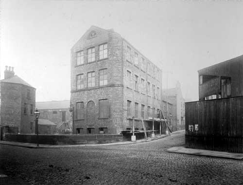 View looking from East Street towards Garden Street, where an unnamed bridge carries the road over Timble Beck. In the centre is a lodging house, 'Queen Lodging House for Men'. Scaffolding is to the right of the building. On the right is Hudson and Co., timber merchants and sawmill. This was to be included in the East Street improvement area. Pictured in March 1910.