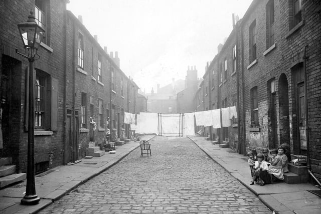 The west side of Cannon Street, which was intersected by Regent Street into two halves. Residents are pictured in the doorways of back to back terrace houses. A line of washing hanging across street, wooden armchair in road. On right, group of children playing on doorstep, with toy bed and doll.