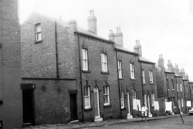 A row of three double fronted back-to-back terraced houses flanked by shared outside toilet yards. A woman outside number 12 hangs clothes on a line stretched across the street. Pictured in June 1973.