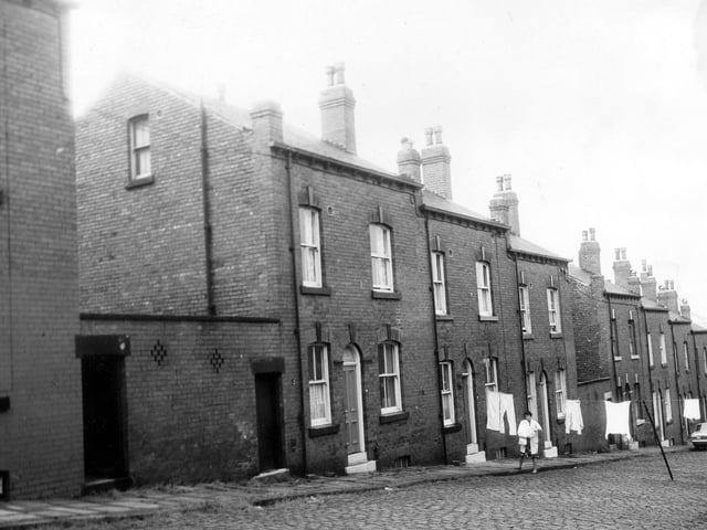 A row of three double fronted back-to-back terraced houses flanked by shared outside toilet yards. A woman outside number 12 hangs clothes on a line stretched across the street. Pictured in June 1973.
