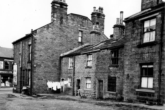Butler Place in August 1963. On the left of the image shops on Town Street are visible. A coach built pram stands by the side wall of number 27 Butler Place at the entrance to Butler Place. On the right are a row of three back-to-back terraced houses numbers 1 to 5 Butler Place. Clothes hang on a line stretched across the front of number 1 while a young girl stands by a tricycle outside number 3. Number 5 on the right has become derelict.