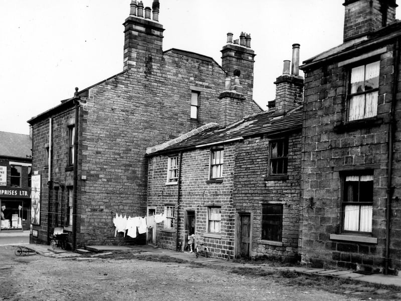 Butler Place in August 1963. On the left of the image shops on Town Street are visible. A coach built pram stands by the side wall of number 27 Butler Place at the entrance to Butler Place. On the right are a row of three back-to-back terraced houses numbers 1 to 5 Butler Place. Clothes hang on a line stretched across the front of number 1 while a young girl stands by a tricycle outside number 3. Number 5 on the right has become derelict.