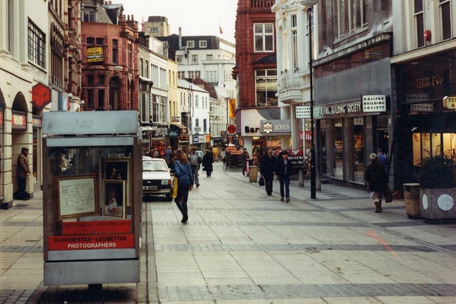 Commercial Street looking in the direction of Briggate in September 1982. On the left is W.H. Smith, formally at number 15 Commercial Street and now in Lands Lane around the corner. Lands Lane cuts across from left to right in the centre in the centre of the image. Barratts shoe shop can be glimpsed right of centre at numbers 36 to 38 Commercial Street. At the right edge is Terry & Co. and then the Halifax Building Society at number 32. In the foreground there is street advertising for Dorchester - Ledbetter Photographers.