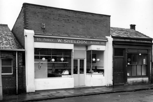 William Sheldon's Family Butchers on Hall Lane in May 1965. A piece of meat hangs in the window, the rest of the stock is likely to be in refrigerators elsewhere in the shop. One the right is  a fish & chip shop, the business of a Mrs Florence Gabbott. An oval sticker in the window shows that this shop is a member of the Leeds & District Fish Fryers Association.