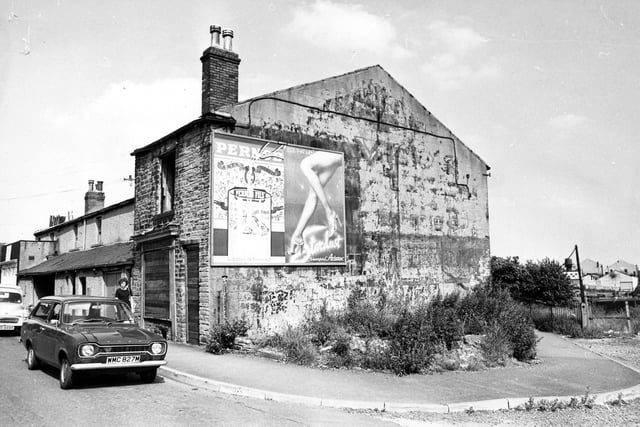 Wortley Road in July 1980 showing No.23-33. No.33 in the foreground is boarded up and has advertisements for Pernod and Aristoc tights on the side. Cars are parked on the road. On the right is Station Road, which formerly led to Armley Moor Station but is now fenced off.