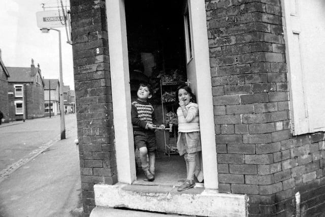 Two children are seen standing in the doorway of a red brick corner shop on Alexandra Road. Inside crisps are on display including a variety called 'Chopstix' and another make called Tudor. On the other side of the road red brick streets run at right angles; from the left are Kelsall Road, Kelsall Place, Kelsall Avenue and Kelsall Terrace.