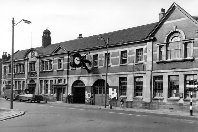 Public Library on the left and the Police Station on the right, both opened on July 24, 1903. On the left on the vehicle entrance is a door marked Juvenile Entrance with vans parked on the left. On the far right stands a double traffic light painted in black and white stripes. On the side of the building in the centre a large ornate clock proclaims the time as 10.14am while people walk below. The road in the foreground is Moor Road, with Hunslet Hall Road at the right of the Police Station. Opposite these buildings is St. Peter's Church. Pictured in August 1964.