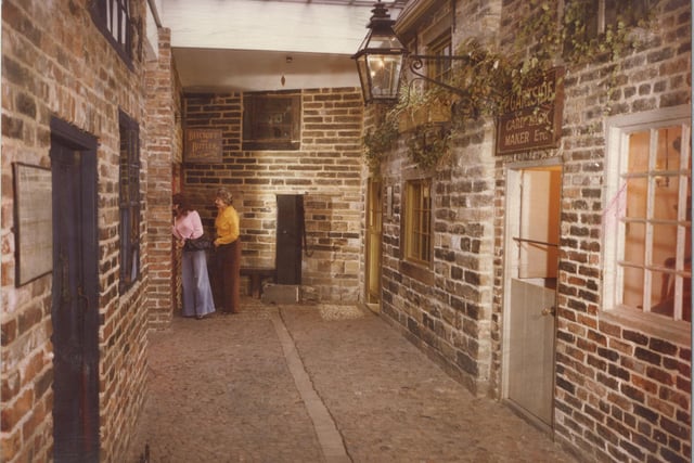The cobbled street of Abbey Fold, part of the Victorian Street replicas at Abbey House Museum. Two women peer into the shop of Beecroft and Butler, blacksmiths, on the left. P. Gartside, card tack maker, etc., sits on the right.