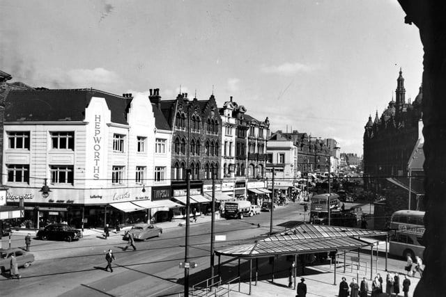 View of New Market Street and onwards to Vicar Lane from the roof of the Corn Exchange in August 1956. At the left edge Central Road joins New Market Street and the Hepworths building stands on the corner at numbers 16 to 18 New Market Street. Timpson's shoe shop is visible at number 14 and Gallons Ltd., Grocer at number 8. Opposite Central Road in front of the Corn Exchange people can be seen waiting for buses near the shelter. The dark shape of Kirkgate Market looms in the background, right, at the junction with Kirkgate. The main road through to North Street then becomes Vicar Lane.