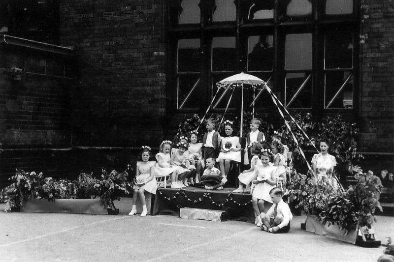 May Day celebrations at Sheepscar Council School in May 1953. The May Queen is seated on a dais, beneath a decorated parasol, surrounded by her attendants. The girls are wearing white dresses, ankle socks and white shoes and have wreaths of flowers in their hair. The boys are smartly dressed, two in waistcoats and one bearing a cushion for the May Queen's crown.