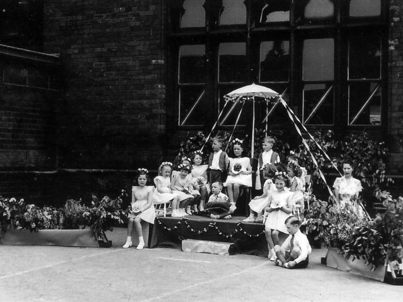 May Day celebrations at Sheepscar Council School in May 1953. The May Queen is seated on a dais, beneath a decorated parasol, surrounded by her attendants. The girls are wearing white dresses, ankle socks and white shoes and have wreaths of flowers in their hair. The boys are smartly dressed, two in waistcoats and one bearing a cushion for the May Queen's crown.