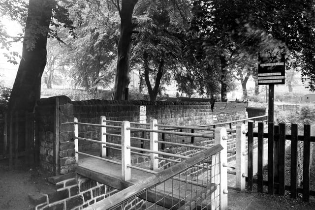 The bridge over Meanwood Beck pictured in June 1948. The concrete footbridge crosses the beck as it flows through a stone channel between Highbury Cricket Club's pitch on the right, and Meanwood Park on the left (over the stone wall). A sign advertising a match between Highbury Works C.C. and Calverley is visible.