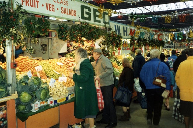 Kirkgate Market during the Christmas season. The stalls are laden with seasonal fresh fruit and vegetables and decorated with hanging holly wreaths. Colourful Christmas decorations are strung across the aisles. The image is thought to date from 1998.