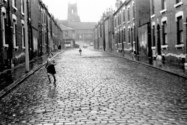 A child runs down Kelsall Grove, a cobbled street of red brick terraced houses. Alexandra Road runs across at the top of the street, with Alexandra Grove leading up from this. The square tower of All Hallows Church in Regent Terrace looms in the background.