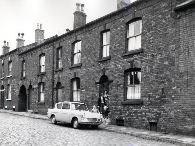 Several people stand in the doorway of a house on Main Street where a car is parked outside. Included in slum clearance plans for the Tong Road area of Armley.