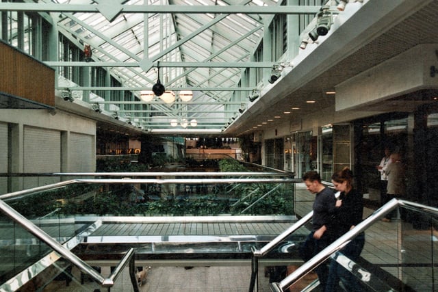 A view along the upper floor of the Centre with a couple in the foreground walking down the stairs. Wade House is partially visible on the right and Morrisons can be seen in the background. Metal beams stretch across the roof with lights and security cameras. Glass panels line the lanes with artificial plants on either side.