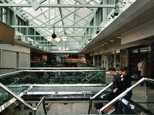 A view along the upper floor of the Centre with a couple in the foreground walking down the stairs. Wade House is partially visible on the right and Morrisons can be seen in the background. Metal beams stretch across the roof with lights and security cameras. Glass panels line the lanes with artificial plants on either side.