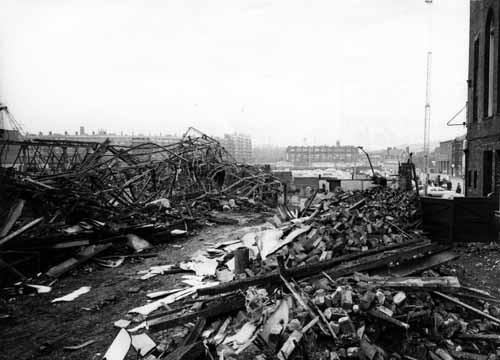 The debris of what had been South Row shops, part of Kirkgate Market at the bottom of Harper Street. This is the aftermath of the fire which had raged through the market on December 13, 1975. The market traders suffered considerably with the loss of their Christmas takings although other temporary sites were found for their stalls. Prince Charles made a visit to the site on the 17th December to help boost the morale of the traders. New York Street is seen on the right with the Central Bus Station and Quarry Hill Flats in the background of the image.