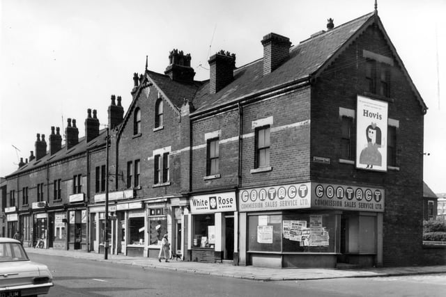A view down Dewsbury Road away from Hunslet Hall Road in August 1964. Shops on the right include Lucille's drapers at number 176, Jones of Oakwood Electrical Appliance dealers at number 172. At number 170 is a wallpaper store with the White Rose Cleaners & Dyers on the right at number 168. On the corner is the empty shop of what was once Contact Commission Sales Service Ltd, now with posters stuck to windows. On the wall of Roxburgh Road an advertising hording promoting Hovis bread. A woman in a head scarf walks a dog past the shops.