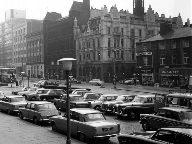 The Headrow from the Town Hall steps showing the junction with East Parade and Calverley Street in June 1967. Cars are parked in front of the Town Hall. Buildings on The Headrow include Pearl Assurance, Yorkshire Assurance, Eagle Star Assurance, a chemist and a tobacconists.