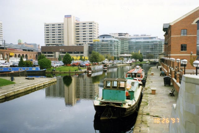 Canal Wharf at the Canal Basin looking in the direction of Victoria Bridge. Two barges are moored in front of Quayside House, right, opposite the Canal Basin, left. The boat in the foreground is The Horbury and acts as storage and supplies for the Kirkstall Flyboat moored behind it. The Kirkstall Flyboat is a purpose-built pleasure boat run by Yorkshire Hire Cruisers Ltd. It is equipped to take private parties of up to 50 people on cruises along the canal and River Aire. On the left a train can be seen crossing over the dark arches, about to enter the City Station. The large building in the background left is the Hilton Hotel in Neville Street.